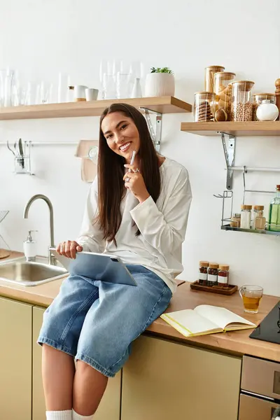 A young beautiful woman sits comfortably in her kitchen, smiling and using her tablet. — Stock Photo