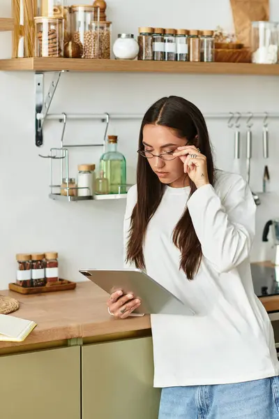 Una giovane donna con i capelli lunghi si trova al suo bancone della cucina, godendo il suo tablet. — Foto stock