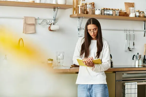 Woman with long hair stands in a bright kitchen, focused on writing notes in her notebook. — Stock Photo