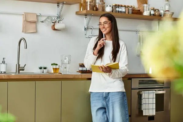 A young woman stands in her cozy kitchen, thoughtfully writing in a notebook with a smile. — Stock Photo