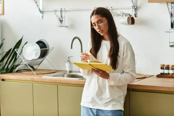 A young woman relaxes in her cozy kitchen, jotting notes in a bright yellow notebook. — стоковое фото