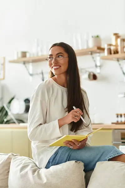 A beautiful young woman relaxes on her couch, writing in a notepad with a smile on her face. — Stock Photo