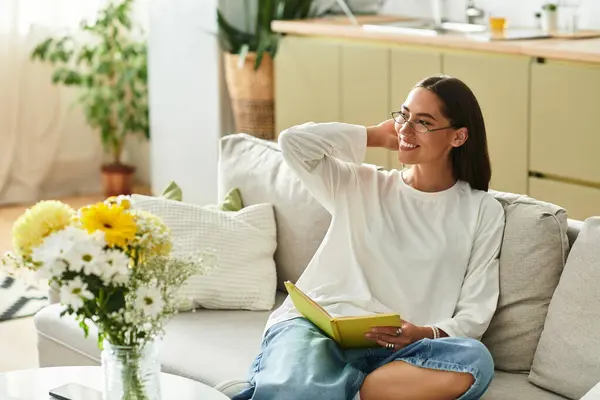 Young woman relaxes on the sofa, reading a book and surrounded by flowers in a cozy home. — Stock Photo