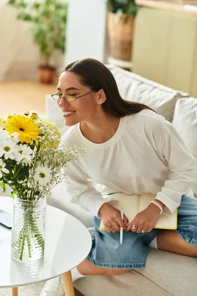 Hermosa mujer se sienta cómodamente en casa, sonriendo mientras admira las flores frescas y escribe. - foto de stock