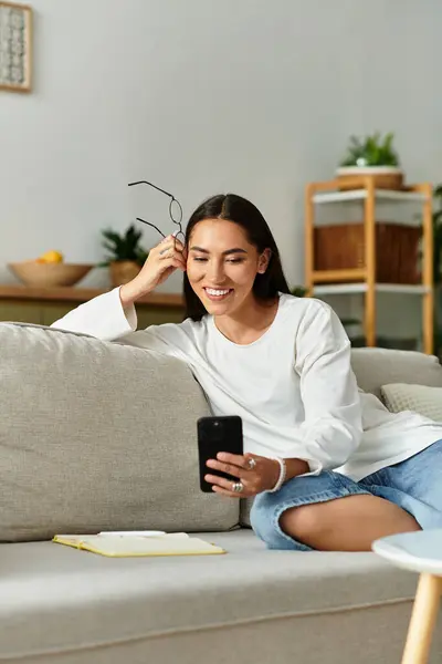Young woman relaxes on a comfy couch, smiling as she interacts with her smartphone at home. — Stock Photo