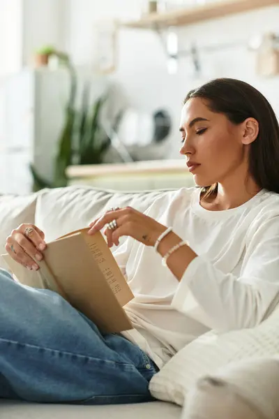 Young woman sits comfortably on the couch, engrossed in reading a captivating book at home. — Stock Photo
