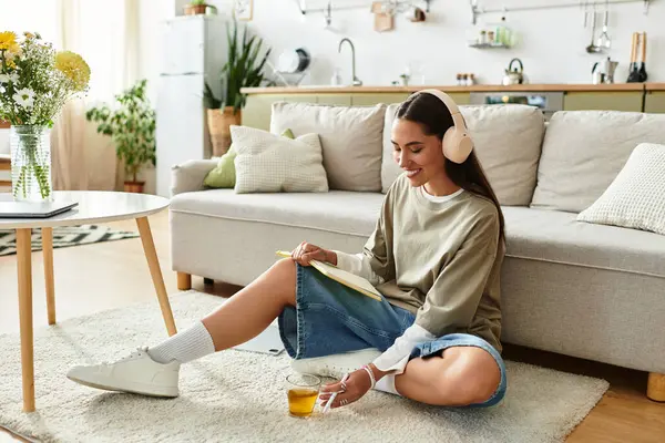 A beautiful young woman sits on the floor, sipping warm tea and writing in her notebook at home. — Stock Photo