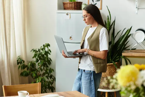 A young woman stands in her home kitchen, working on her laptop while surrounded by plants. — Stock Photo
