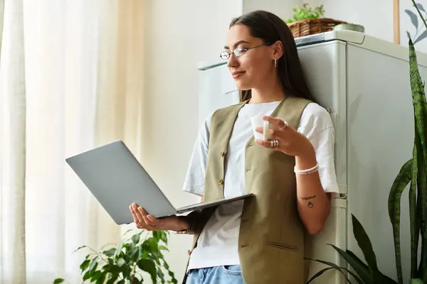 A young woman relaxes at home, holding a coffee cup and working on her laptop in a cozy space. — Stock Photo