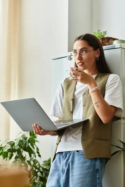 Relaxed young woman stands at home, savoring tea and focusing on her laptop work. — Stock Photo
