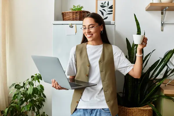 A young woman works on her laptop at home, joyfully holding a coffee cup amidst plants. — Stock Photo