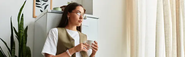 A young woman stands comfortably at home, sipping a warm drink while surrounded by greenery. — Stock Photo