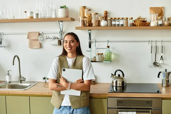 Una hermosa joven sonríe mientras sostiene su tableta en una cómoda cocina llena de cálida decoración. - foto de stock