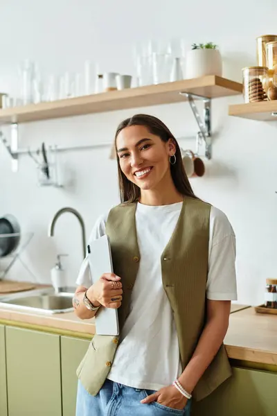 A beautiful young woman smiles while holding a laptop in a charming kitchen filled with light. — Stock Photo