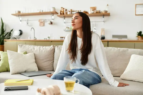 Relaxing at home, a beautiful young woman sits comfortably on the sofa, lost in thought with tea. — Stock Photo