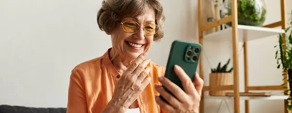 Mujer madura en ropa cómoda disfruta de un momento de alegría mientras usa su teléfono inteligente en casa. — Stock Photo
