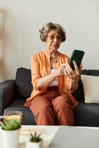 Mature woman sits comfortably on a couch, focused on her smartphone in a warm and cozy home setting. — Stock Photo