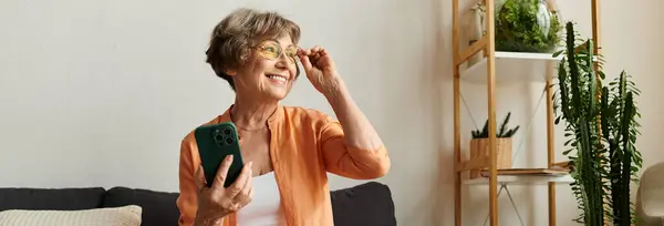 Delighted mature woman in comfortable attire smiles while holding her smartphone at home. — Stock Photo