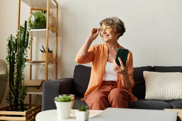 A senior woman smiles at home, comfortably dressed as she interacts with her smartphone. — Stock Photo