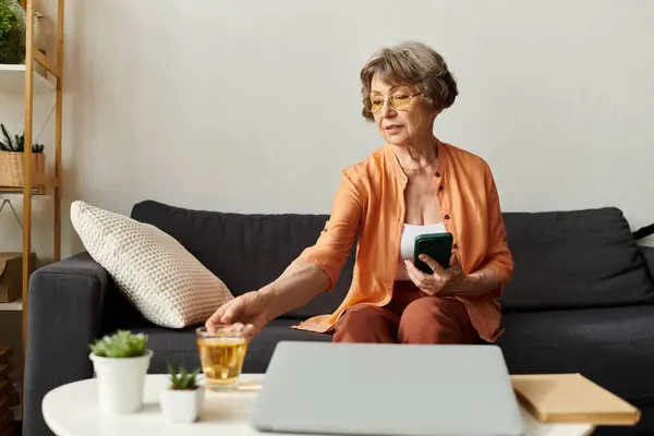 Senior woman in cozy attire sits comfortably at home, sipping tea while reaching for a snack. — Stock Photo
