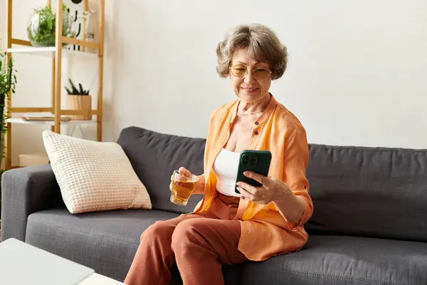 A senior woman in comfy clothes relaxes on a couch, sipping a drink and using her smartphone. — Stock Photo