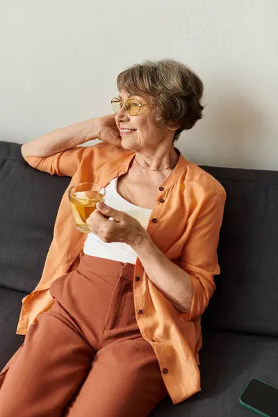A senior woman in cozy attire enjoys a warm drink on the sofa with a smile. — Stock Photo