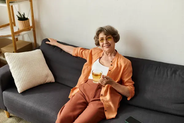A senior woman in cozy clothes lounges comfortably on her sofa, sipping a warm drink and smiling. — Stock Photo