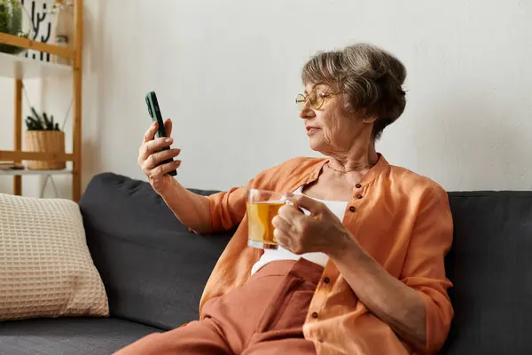 In a comfortable setting, a mature woman relaxes on her couch sipping tea and looking at her phone. — Stock Photo