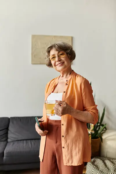 Mujer madura alegremente sosteniendo una bebida mientras se relaja en un ambiente acogedor en casa. - foto de stock