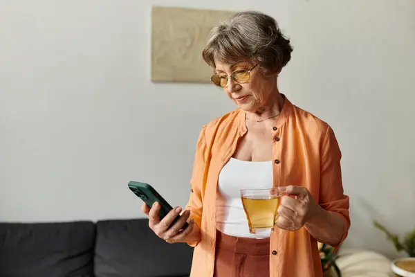 Mujer madura vestida con ropa cómoda se relaja en casa, bebiendo té y usando su teléfono. - foto de stock
