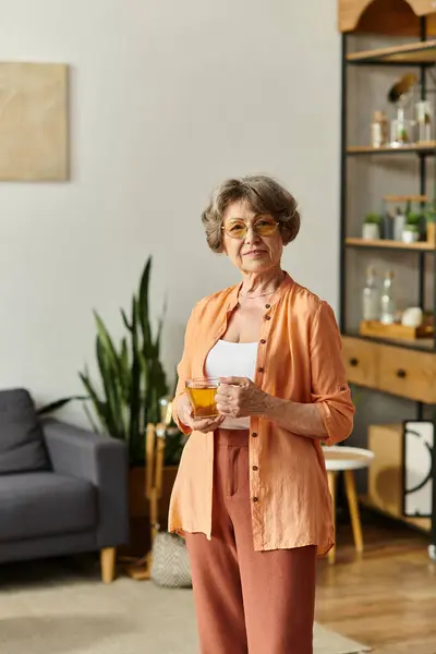 Senior woman enjoys a warm cup of tea while embracing comfort in her stylish living space. — Stock Photo