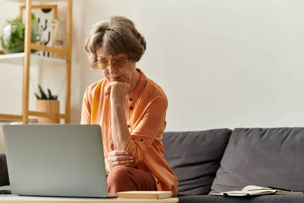 A mature woman in warm attire sits comfortably on a couch, deeply engaged with her laptop screen. — Stock Photo