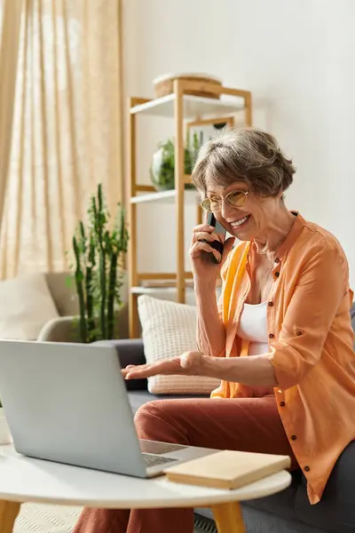 Una mujer mayor en traje acogedor chats felizmente en el teléfono mientras usa su computadora portátil en casa. - foto de stock
