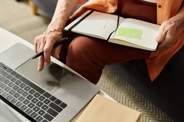 Mature woman in cozy attire focused on her laptop while taking notes in her notebook at home — Stock Photo