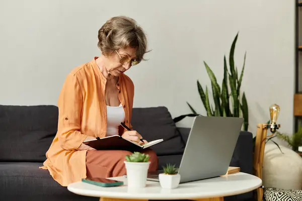 A mature woman sits comfortably at home, engaged in writing notes while her laptop rests nearby. — Stock Photo