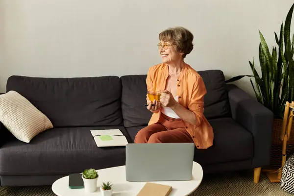 A mature woman in cozy attire sits comfortably on her sofa, smiling while holding a drink. — Stock Photo