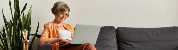 Mature woman in comfy attire smiles as she engages with her laptop in a cozy living space. — Stock Photo
