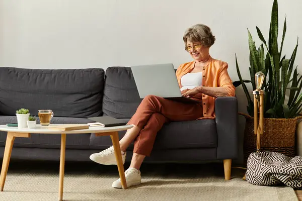 A mature woman relaxes in stylish attire, focusing on her laptop in a comfortable living space. — Stock Photo