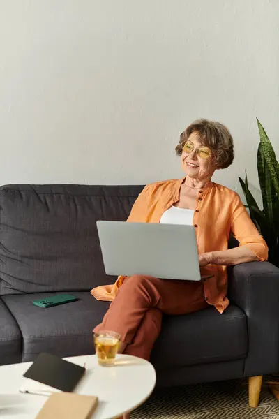 Una mujer mayor con ropa cómoda sonríe en un sofá mientras usa su computadora portátil en una habitación acogedora. - foto de stock