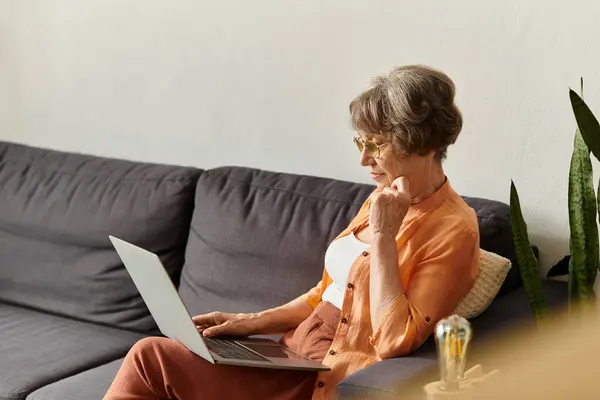 Mature woman relaxes at home in comfy attire, focused on her laptop in a cozy environment. — Stock Photo