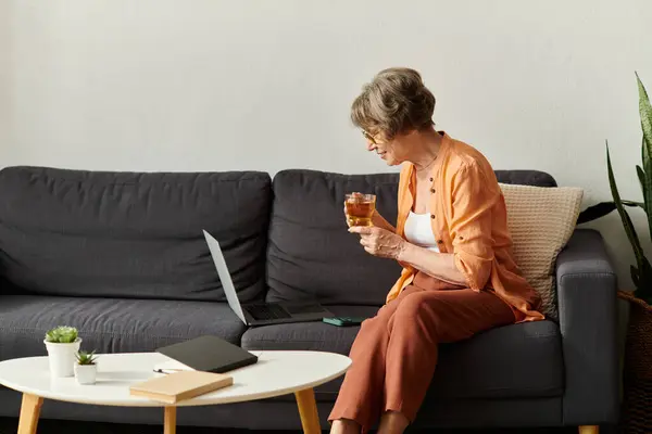 A senior woman enjoys a warm drink on a cozy couch while browsing on her laptop in her home. — Stock Photo