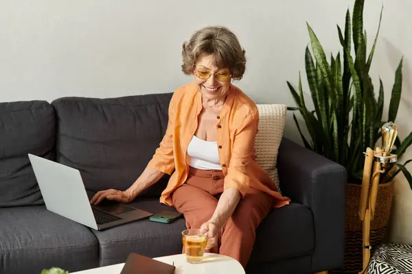 Mature woman sits comfortably at home, sipping tea while engaging with her laptop. — Stock Photo