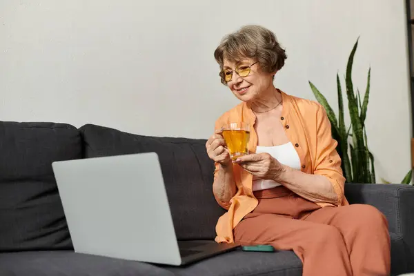 Una mujer madura se relaja en un sofá con una bebida caliente, usando su computadora portátil en un ambiente acogedor. - foto de stock