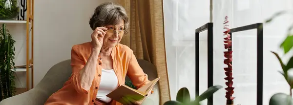 A senior woman in comfortable attire relaxes at home while reading a book by the window. — Stock Photo