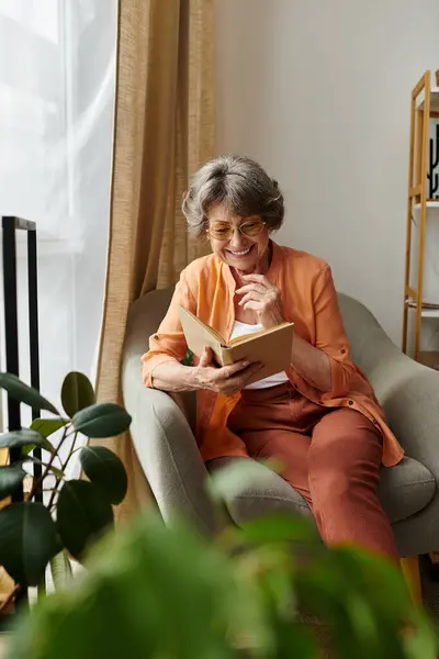 Mujer madura en traje acogedor sonríe mientras lee un libro en un espacio de vida cálido y acogedor. - foto de stock