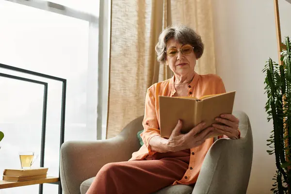 Mature woman relaxes in her home, engrossed in a book, basking in the warm afternoon light. — Stock Photo