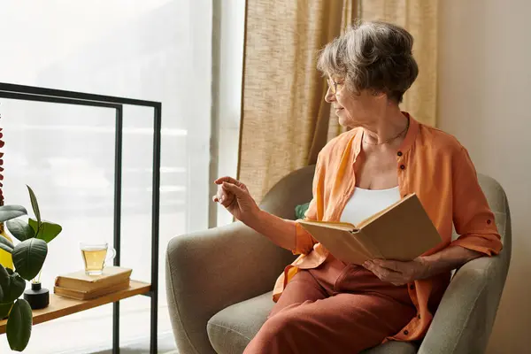 A mature woman relaxes in her comfortable chair while reading a book and savoring a warm beverage. — Stock Photo