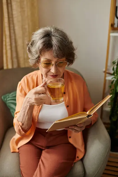 Mature woman relaxes in comfortable attire while sipping tea and reading a book at home. — стоковое фото