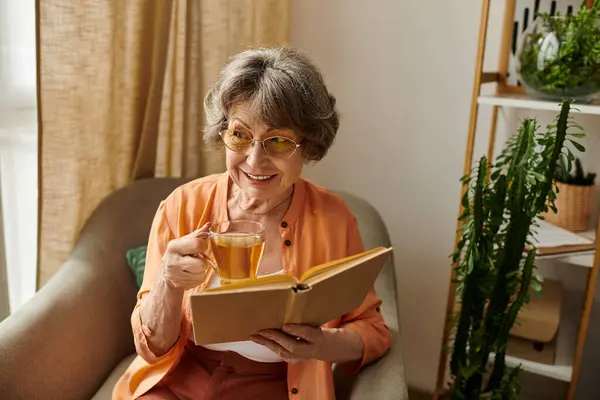 A senior woman enjoys her cozy time at home, sipping tea while reading a book in her chair. — Stock Photo