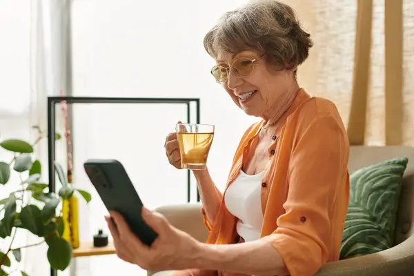 Una mujer mayor se relaja en casa, bebiendo té y sonriendo mientras usa su teléfono inteligente. - foto de stock